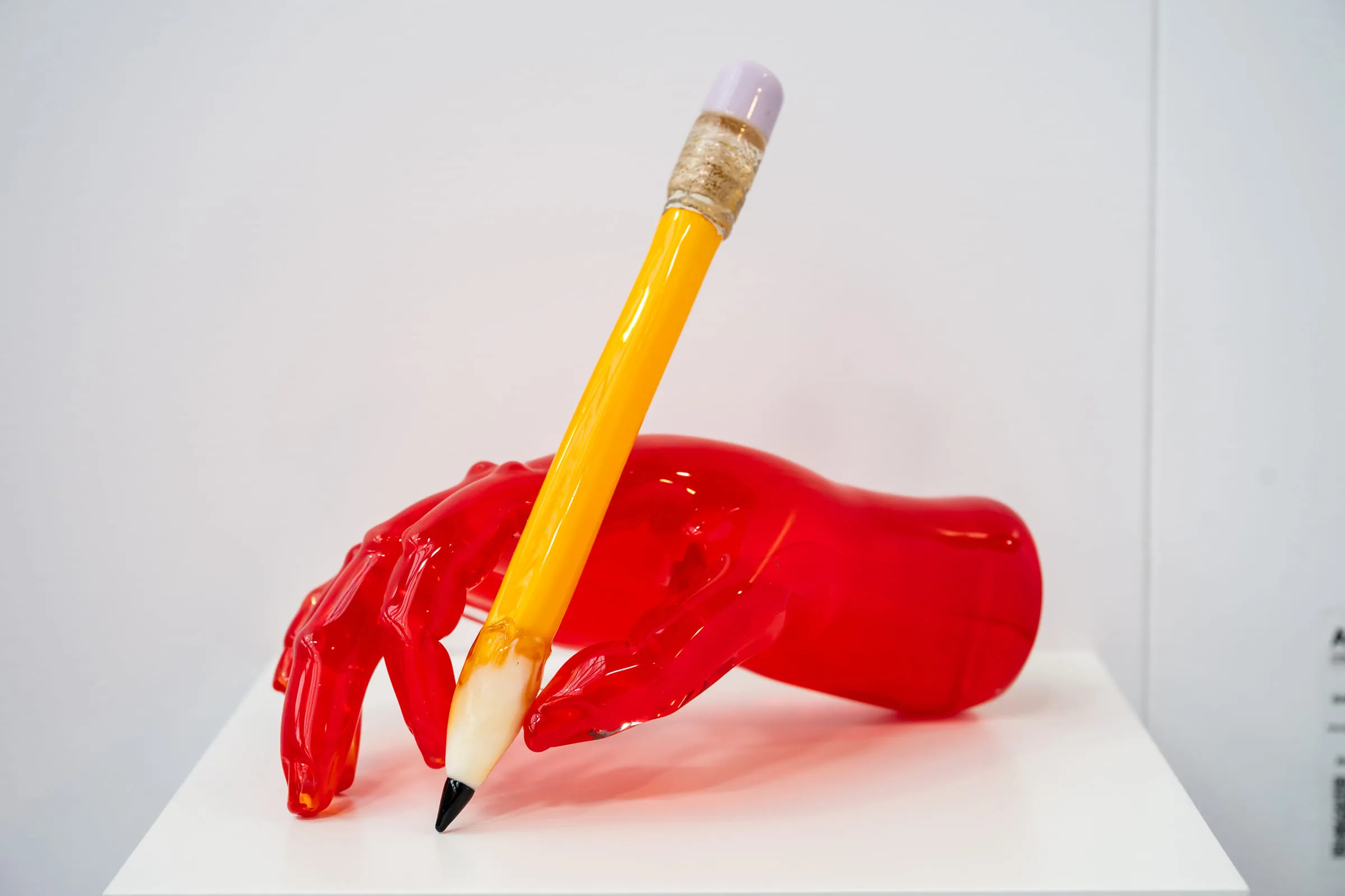 A red glass hand holding an oversized yellow pencil, on a white plinth.