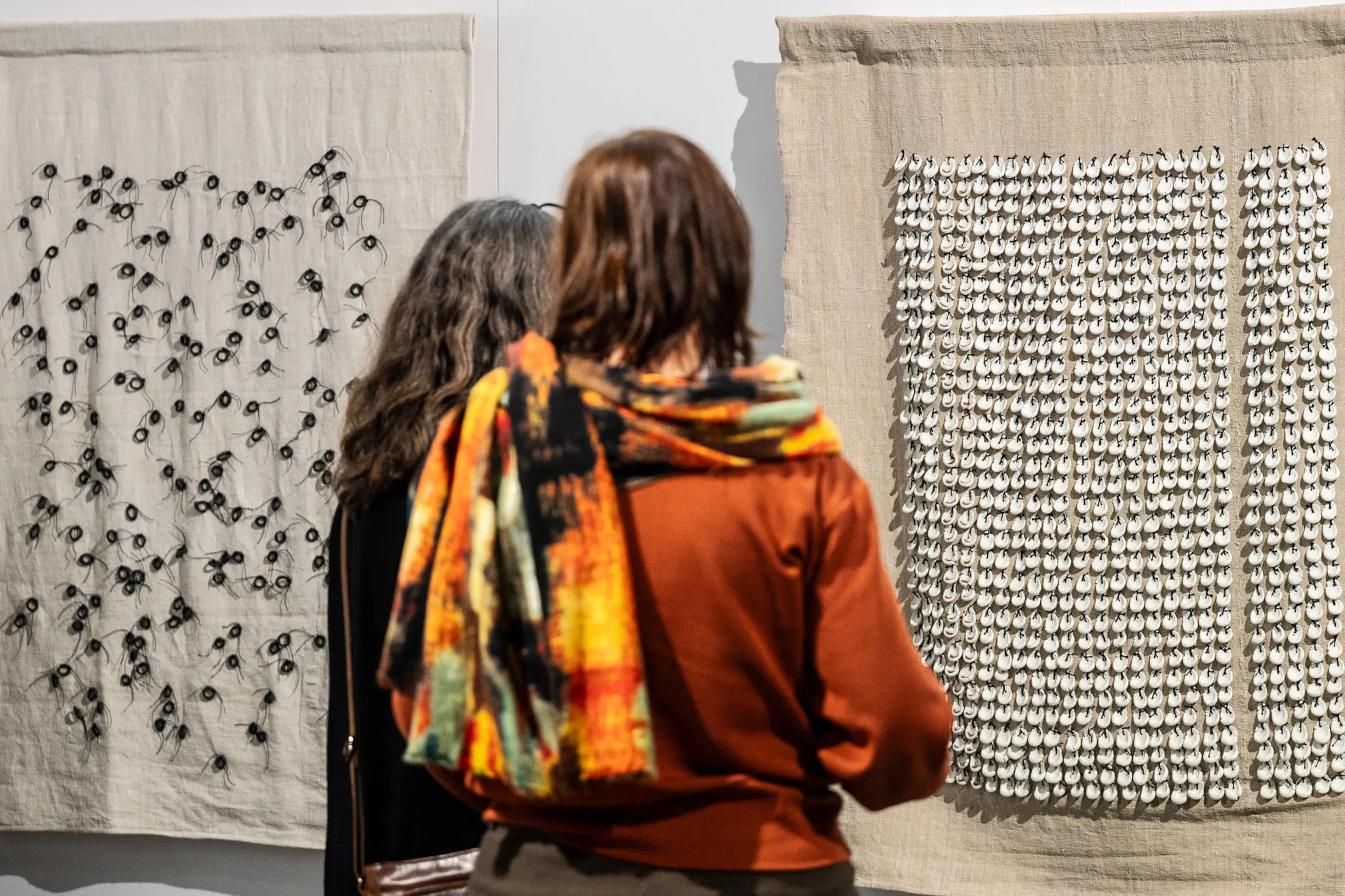 Two visitors in front of two linen hangings covered with small ceramic elements.